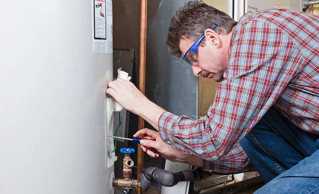 A specialized technician in safety glasses using a screwdriver to repair the heating element of a white residential water heater in a Columbia, SC home.