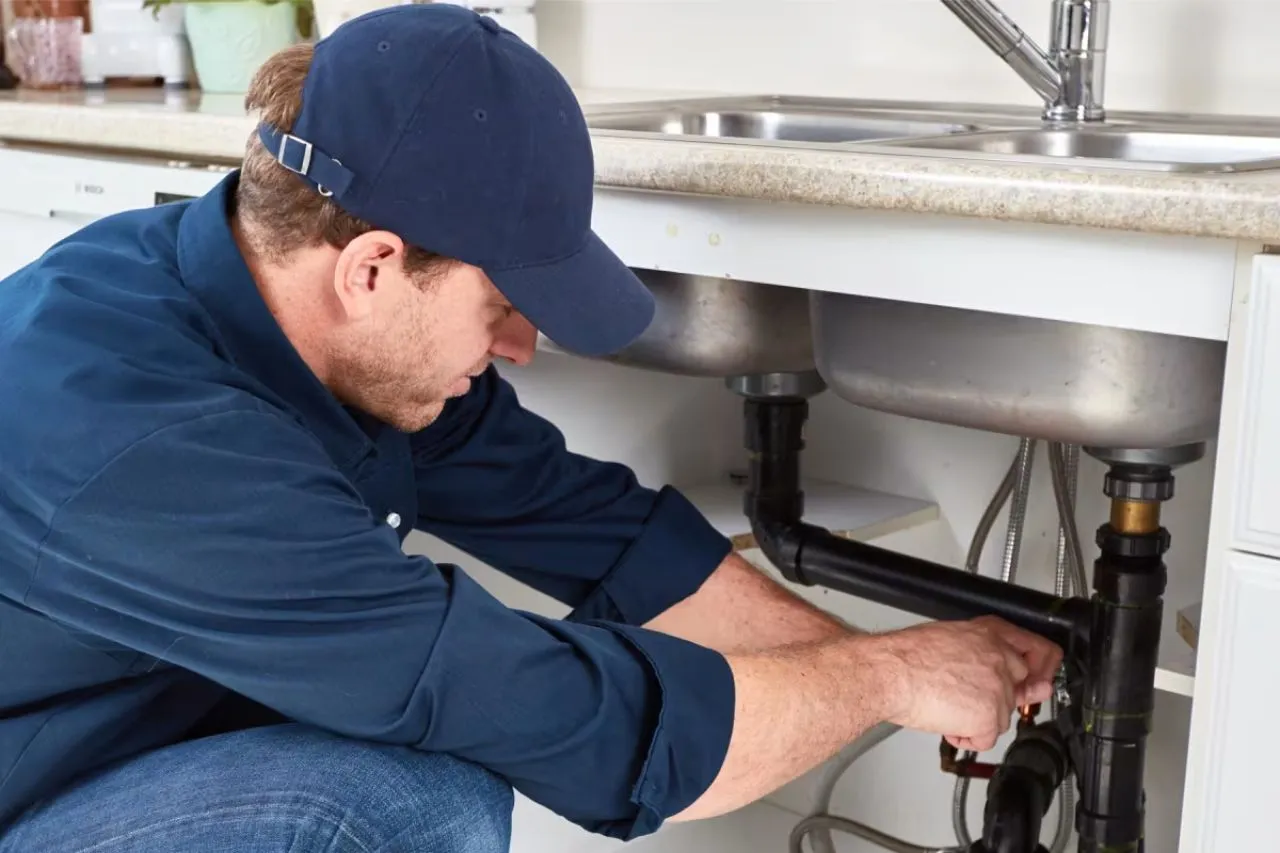 A professional plumber in a blue uniform and cap carefully inspecting and repairing a kitchen sink drainage system in a Columbia home.
